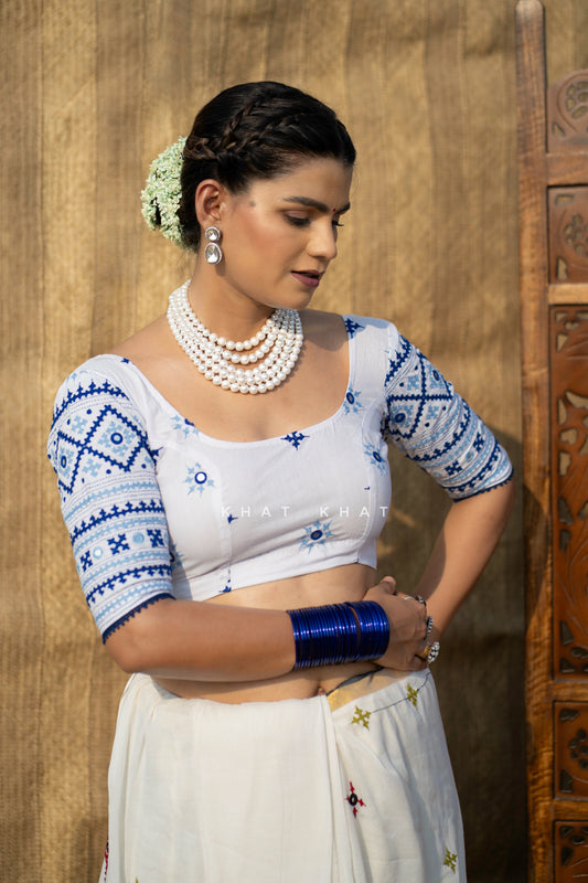 A woman wearing a white and blue kutch embroidered traditional blouse and white skirt, styled with a saree , white pearl necklaces, silver earrings, and blue bangles, standing against a rustic wooden backdrop.