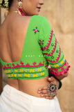 A woman shown from the back wearing a bright green Kutch ahnd embroidered blouse with pink, yellow, and blue decorative patterns and a white skirt. The blouse has a wide neckline and ornate sleeve embroidery with pom-poms.